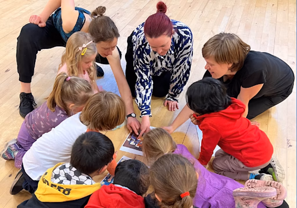 A group of children sit in a circle and point towards something on the floor.