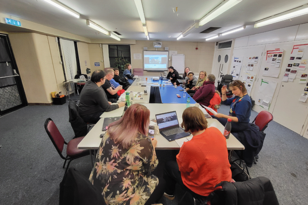 A group of people sit around a large rectangular table in a brightly lit room, engaged in a collaborative meeting or workshop. Laptops, notebooks, drinks, and printed materials are spread across the table. Some participants are looking at a laptop screen, while others are listening or reading documents. On the far wall, a projector displays an image on a screen. The room has white walls, posters, and papers with images and notes taped to the right side.