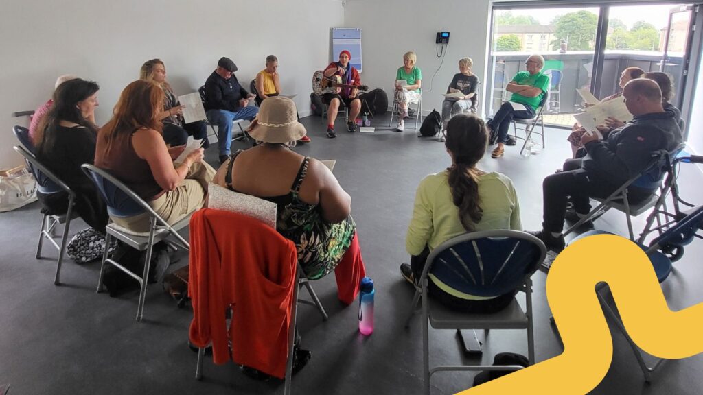 A group of people sit in a circle on folding chairs in a bright room with large windows. They are participating in a singing workshop, holding song sheets, and one person in the centre plays an acoustic guitar. The mood appears relaxed and engaged. A yellow graphic swirl overlays the bottom right corner of the image.
