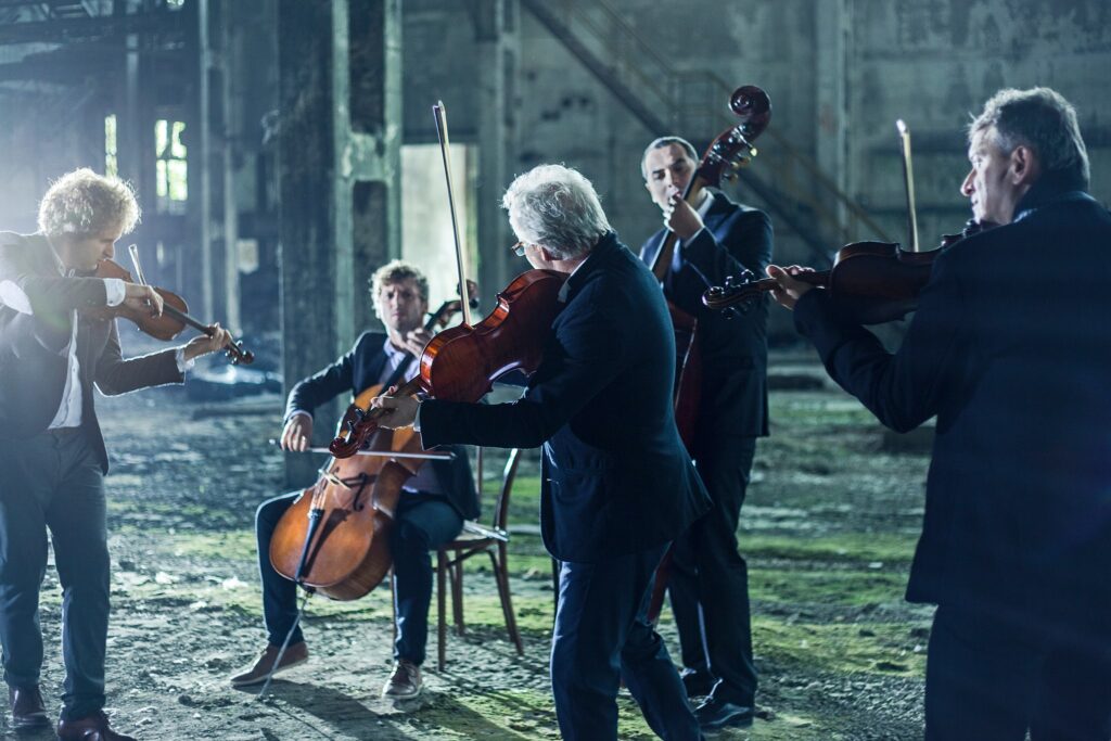 A STRING quintet, five men play different string instruments in a warehouse, all wear formal suits
