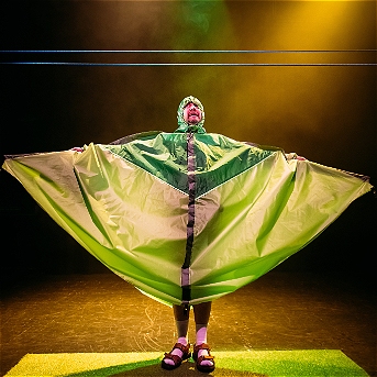 performer stands on stage under dramatic yellow and green lighting, wearing a large, bright green costume that spreads out like wings or a tent. The costume covers their body and has a triangular pattern. The performer is also wearing a hood, purple sandals, and socks, standing on a small patch of artificial grass.