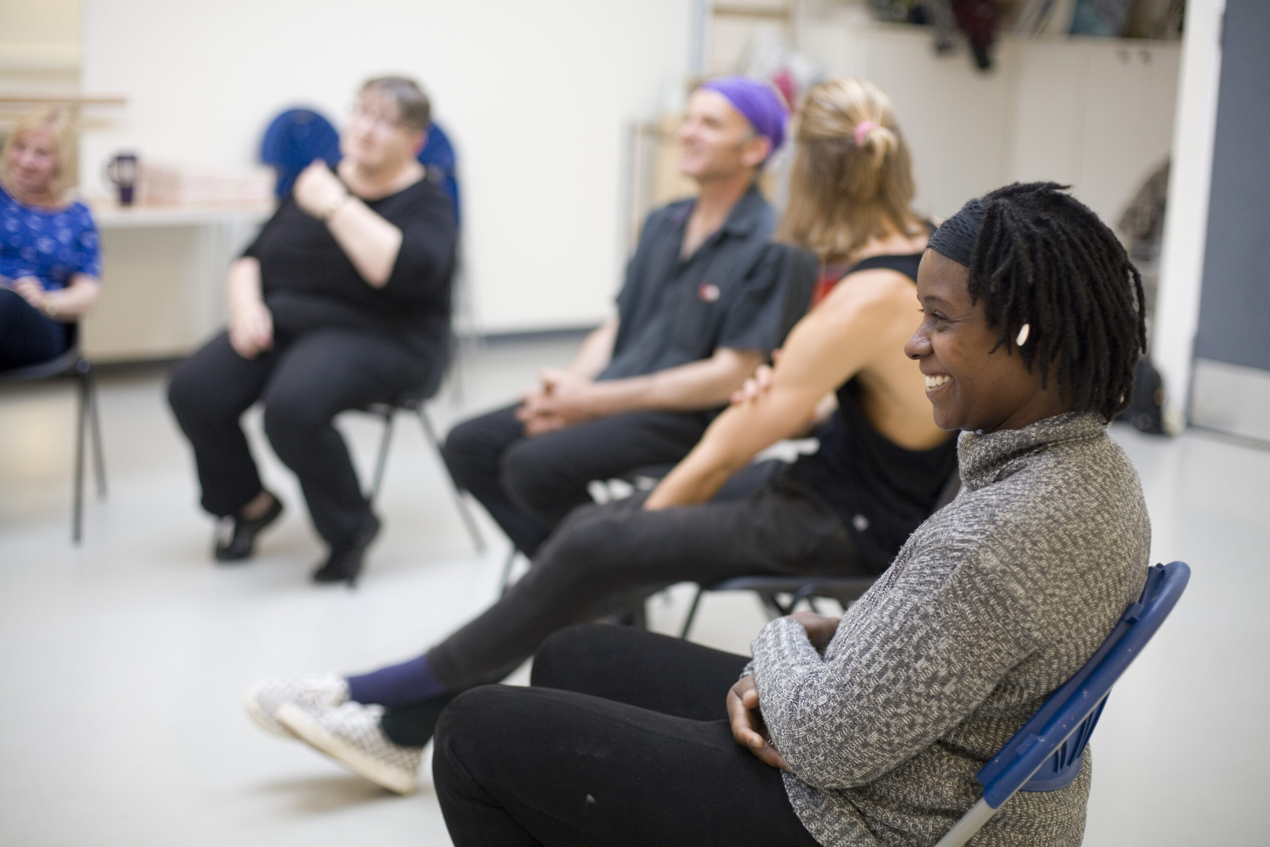 A group of adults sit in a circle on chairs in a bright room, smiling and engaged in conversation during a relaxed workshop rehearsal. The focus is on a person in the foreground wearing a grey sweater, who is smiling warmly. The atmosphere appears friendly and inclusive