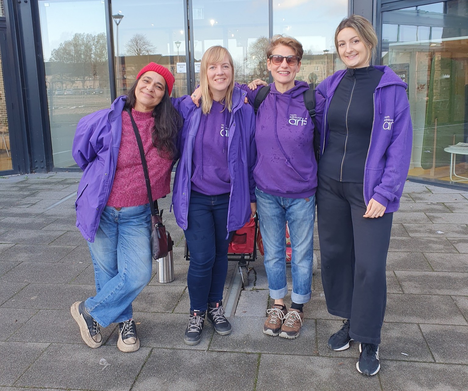 Four women (the Play Rangers team) standing together outside a modern glass building, smiling at the camera. They are wearing purple jackets & hoodies. One person on the left wears a red beanie and pink sweater; the others wear casual outdoor clothing, with two wearing purple hoodies. They look friendly and relaxed, with arms around each other.