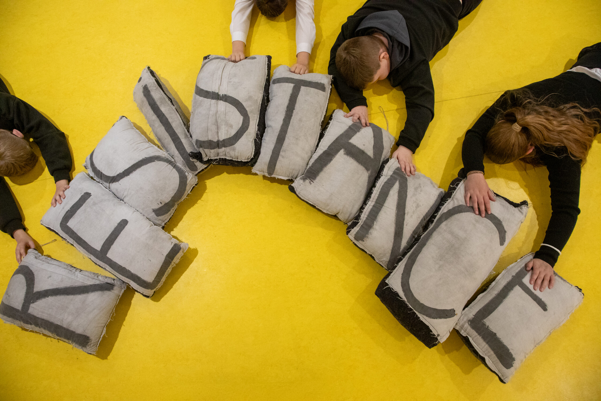 Overhead view of young people lying on a bright yellow floor, arranging grey fabric cushions that spell out the word “RESISTANCE.”