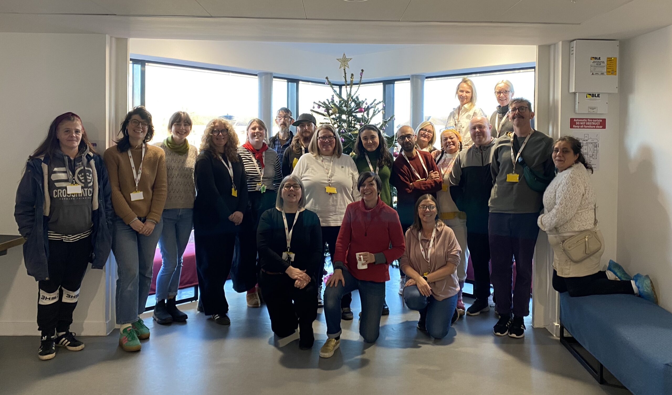 The NEA staff team members pose together in front of a decorated Christmas tree in the NEA building. Some are standing while others kneel in the front row, all smiling at the camera. Many wear lanyards.