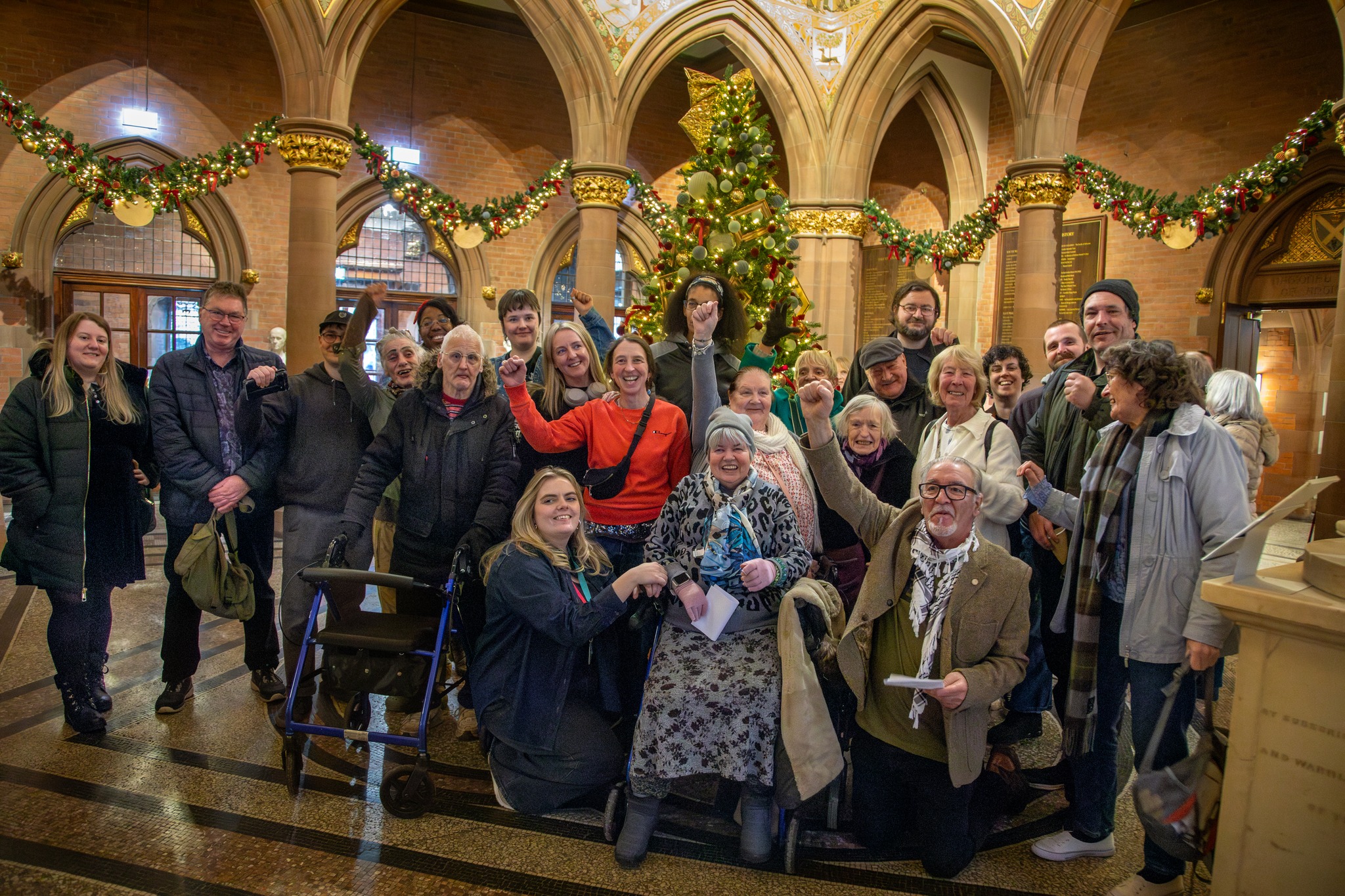 A diverse group of people pose together inside the grand hall of the National Galleries of Scotland: Portrait Gallery, decorated with Christmas garlands and a tall Christmas tree. Many people raise their fists in a gesture of solidarity, smiling and facing the camera. The group includes people of different ages and abilities, gathered closely in a celebratory, collective moment.