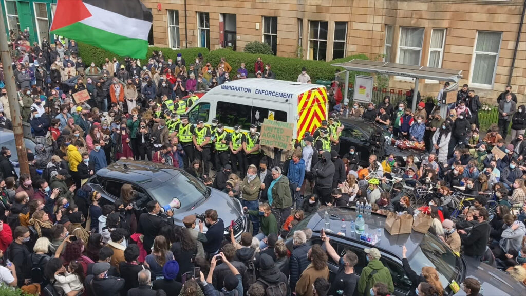 A large crowd of protesters surrounds an Immigration Enforcement van on Kenmure Street, with a line of police officers standing between the vehicle and demonstrators. Many people are sitting or standing close together, some holding signs, others filming with phones. A Palestinian flag is raised above the crowd, and nearby cars are blocked in as the protest fills the street.