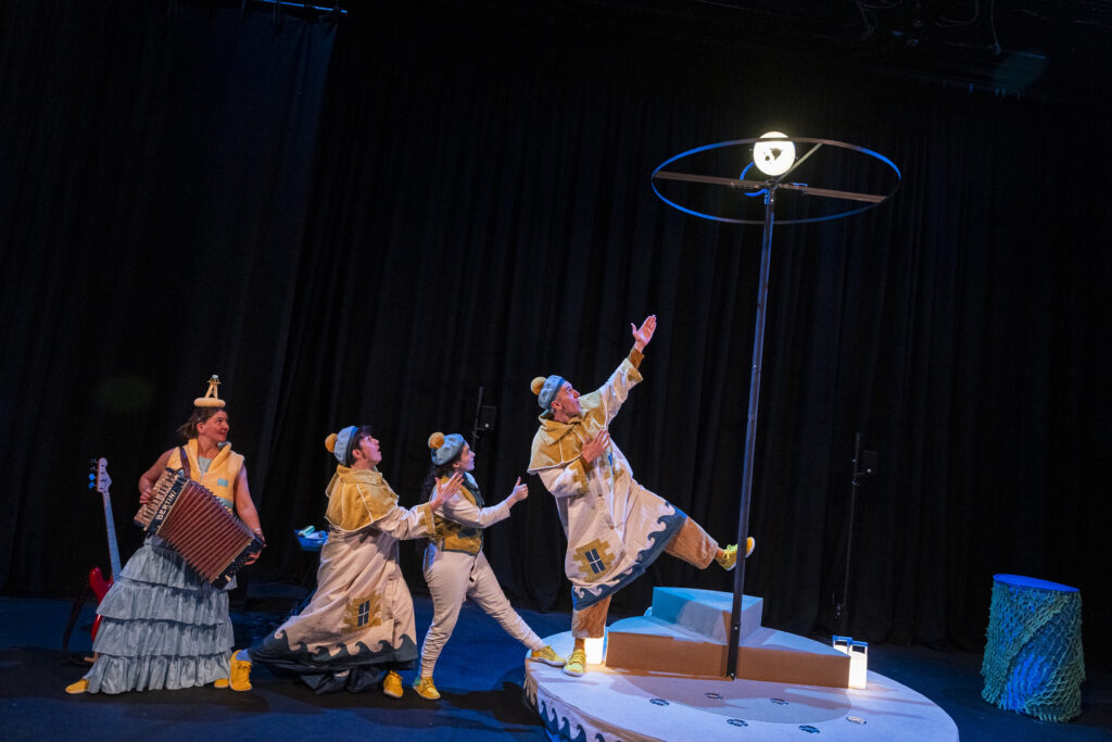 LESS TEXT UP TO 80 WORDS11:24Claude responded: Four performers on a darkened stage.Four performers on a darkened stage. Three stand left — one in a silver ruffled skirt holding an accordion, two in cream and mustard nautical costumes with bobble hats — watching a fourth on a raised white boat-shaped platform, stretching dramatically upward with one arm and leg raised. A circular lighting rig glows above on a tall pole. A red electric guitar leans against the backdrop. All performers wear yellow trainers.