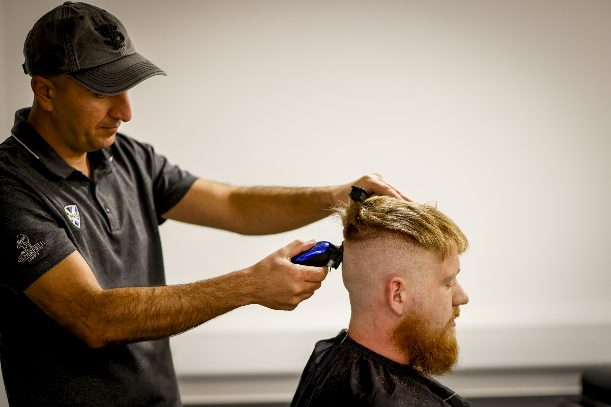 A barber wearing a cap and grey polo shirt uses clippers to trim the hair of a seated client with red hair and a beard, who is wearing a black cutting cape.