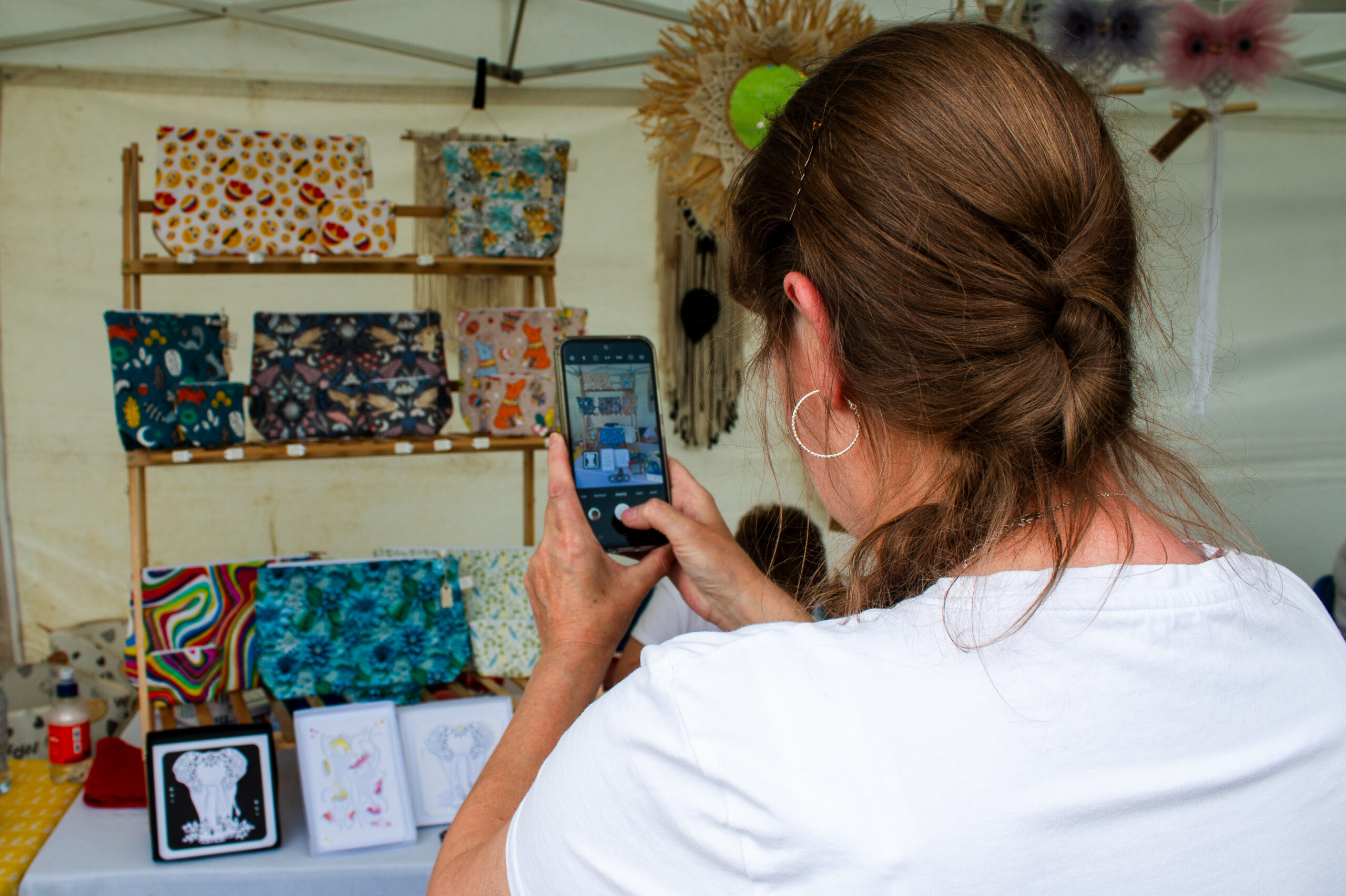 A woman photographs a market stall display of colourful patterned pouches, cards, and handmade crafts on a wooden shelf, using her smartphone.