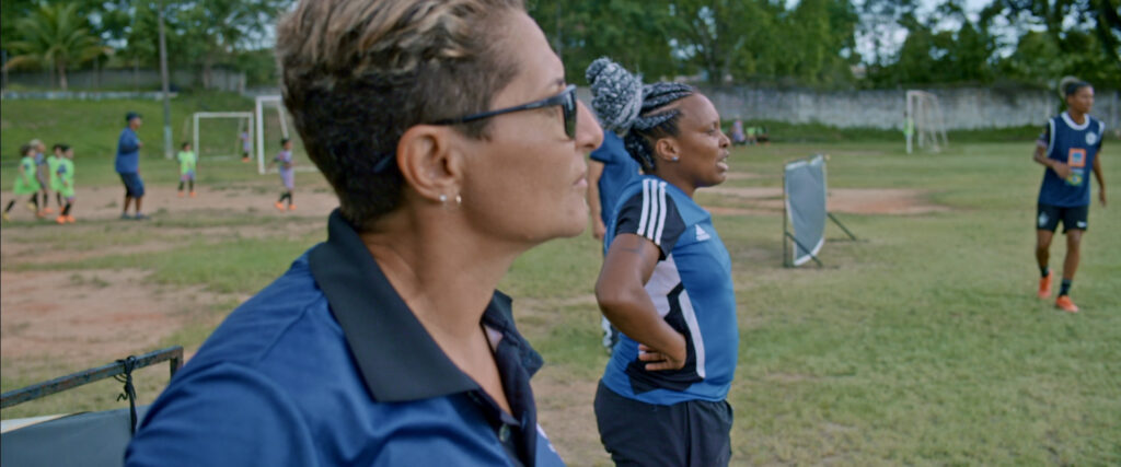 Still from the documentary First Women, showing two women coaches watching a football training session on an outdoor pitch, with young players and a football goal visible in the background
