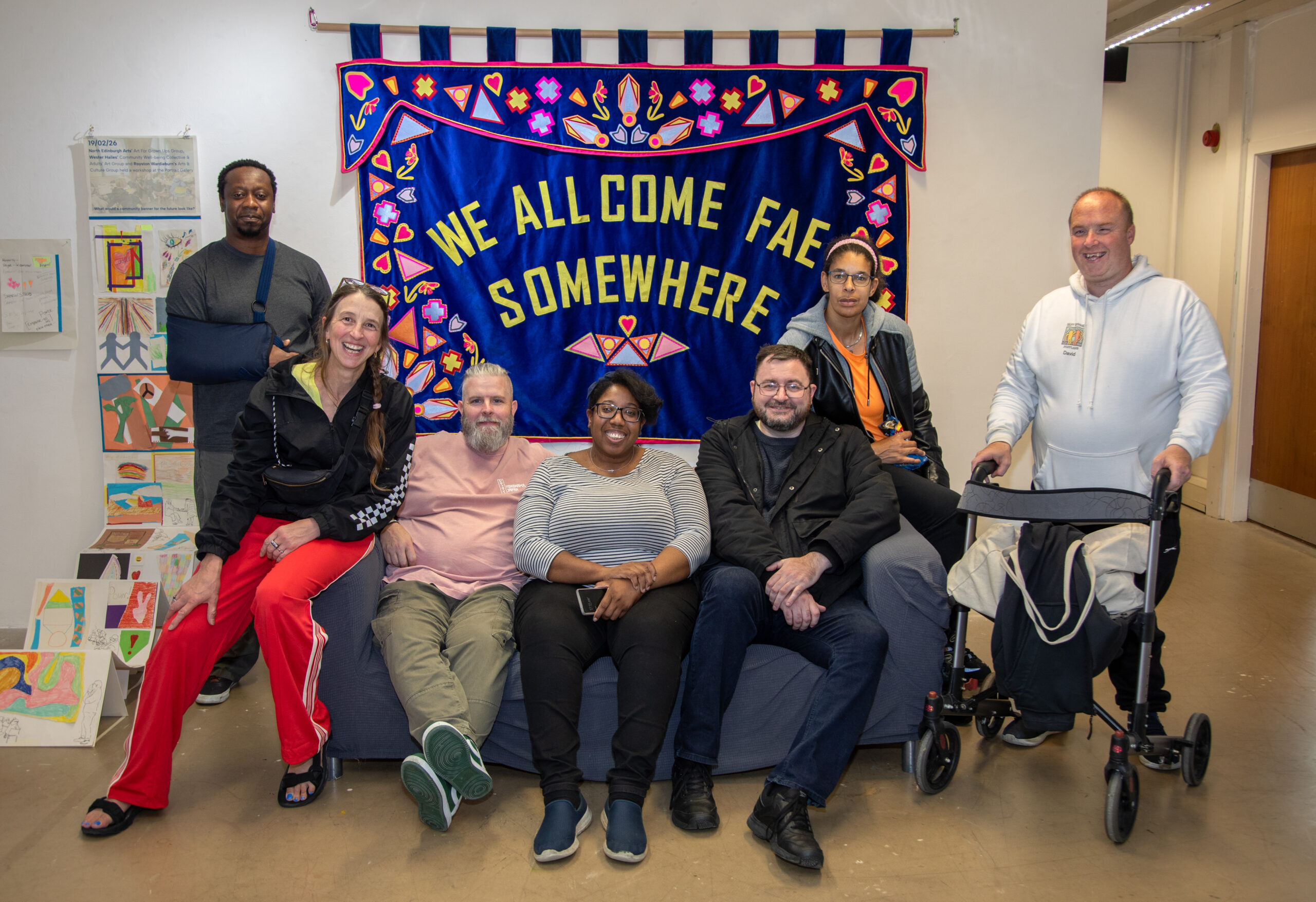 A group of seven smiling people gather around a sofa in a gallery space, in front of a large blue banner reading "We All Come Fae Somewhere," decorated with colourful appliqué hearts, crosses and geometric shapes. Handmade artworks are displayed on a stand to the left