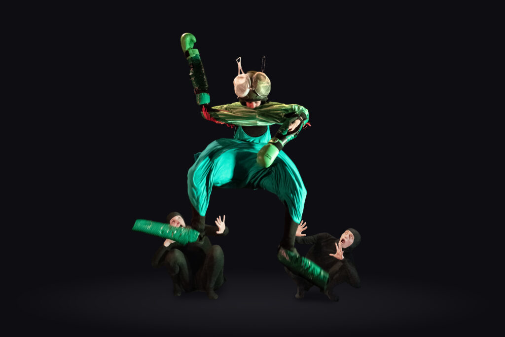 A performer in a green fly costume towers over two startled, crouching performers in black against a dark background.
