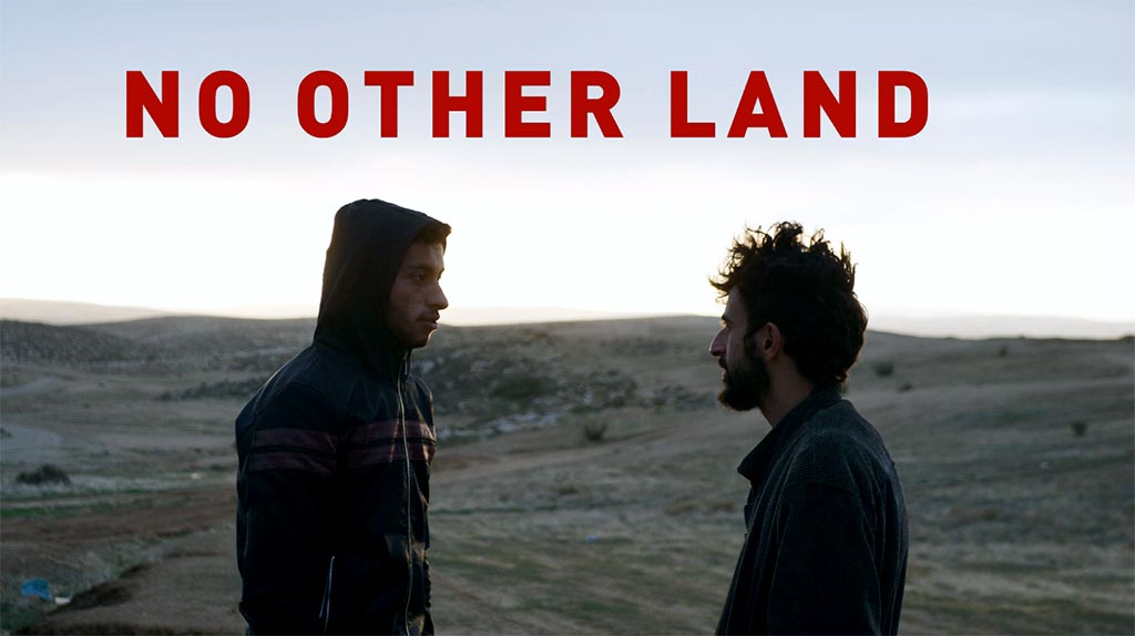 Two men stand facing each other on a barren, hilly landscape at dusk, shown in profile. The man on the left wears a dark hooded jacket with red stripes across the chest. The man on the right has dark curly hair and a beard and wears a dark jacket. The pale sky glows behind them. Red capital letters across the top read “NO OTHER LAND”.
